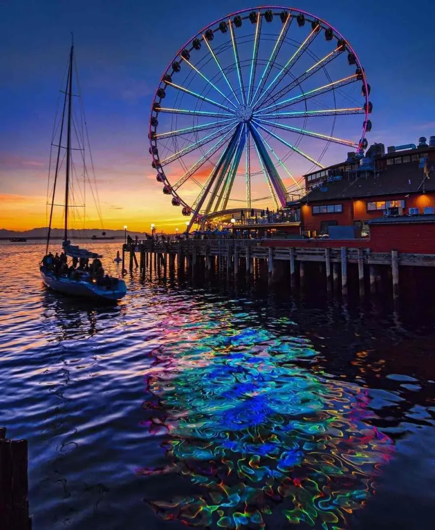 The glowing Seattle Great Wheel reflects in the water at sunset by a sailboat in Seattle in July.