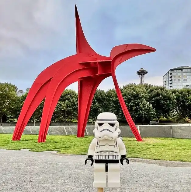 A Lego Stormtrooper stands before a large red sculpture in a grassy downtown Seattle park.